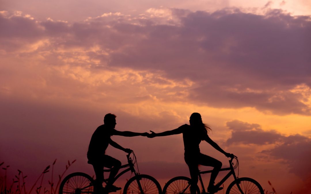 Silhouettes de Deux personnes chacune sur un velo au soleil couchant. Les deux personnes essaient de se tenir la main à vélo. C'est une métaphore de l'infidélité et de la fidélité à soi. Comment rester ensemble